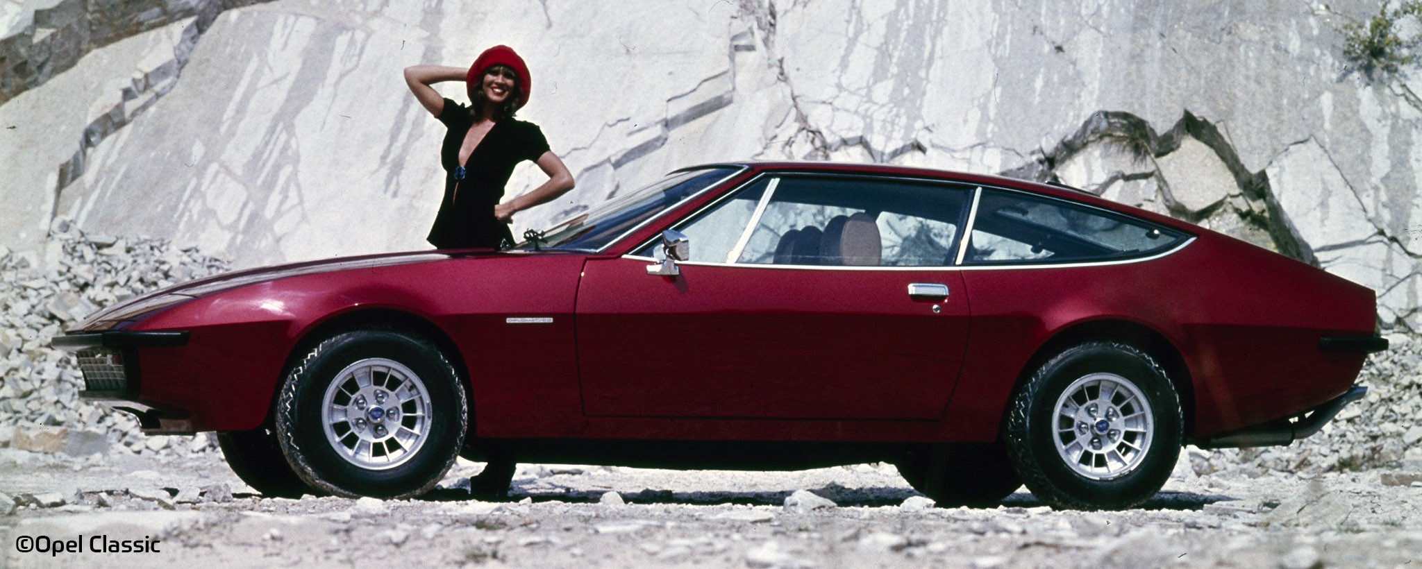 The picture shows an Opel Monza A in front of a stone wall