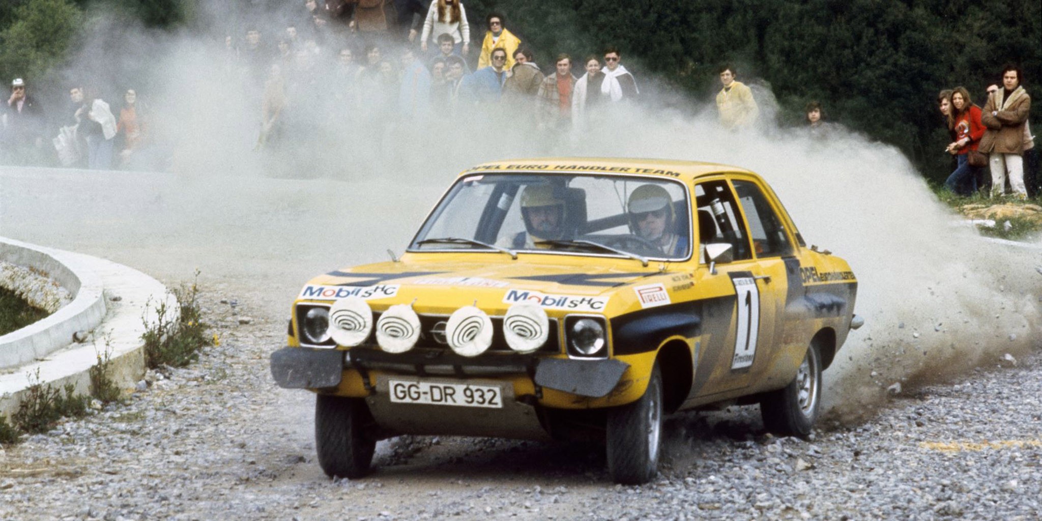 Here, Walter Röhrl in a yellow Opel Ascona A stirs up a lot of dust by taking a right-hand bend during a rally.