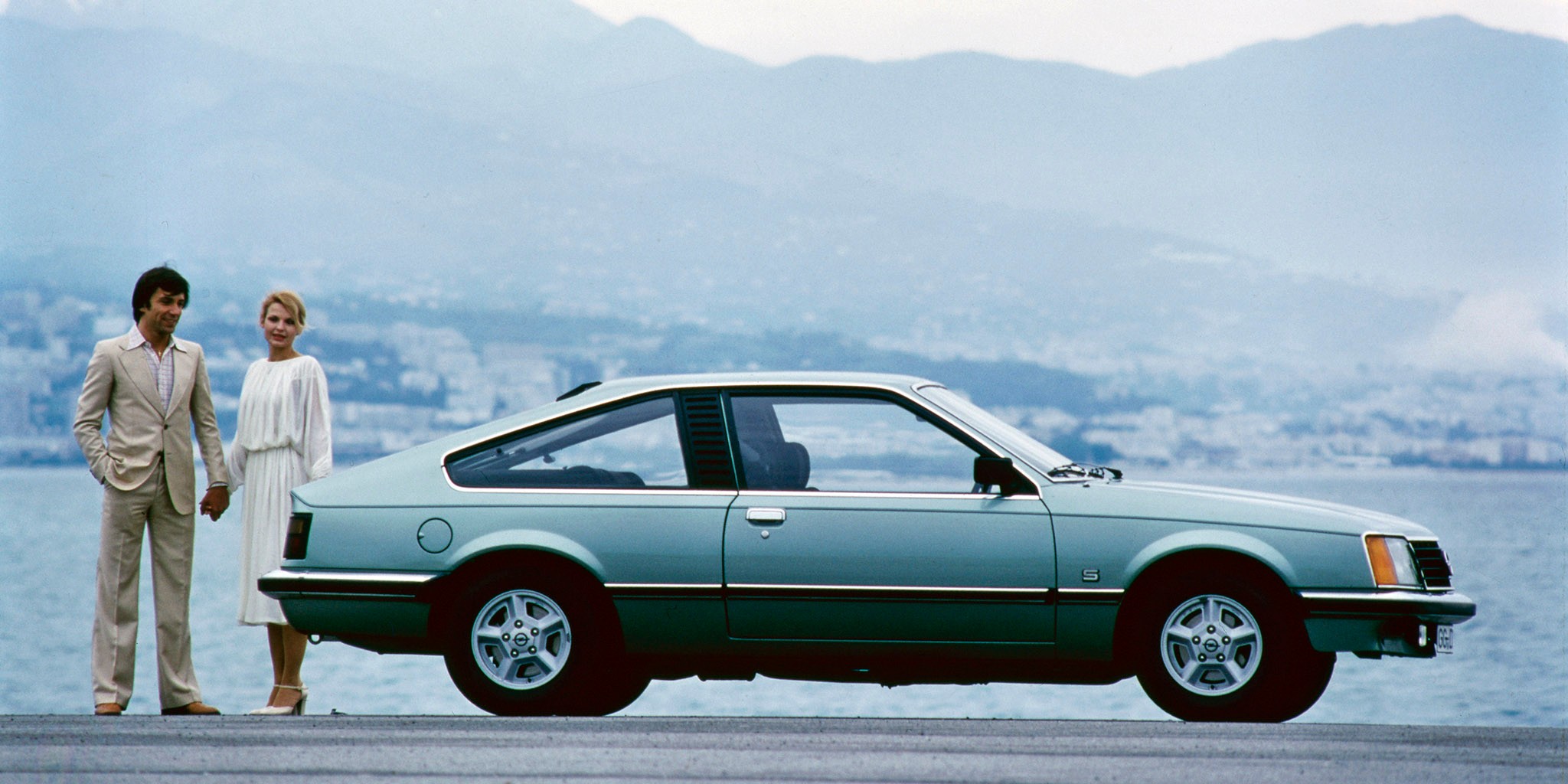 A couple is standing by a lake next to a blue Opel Monza.