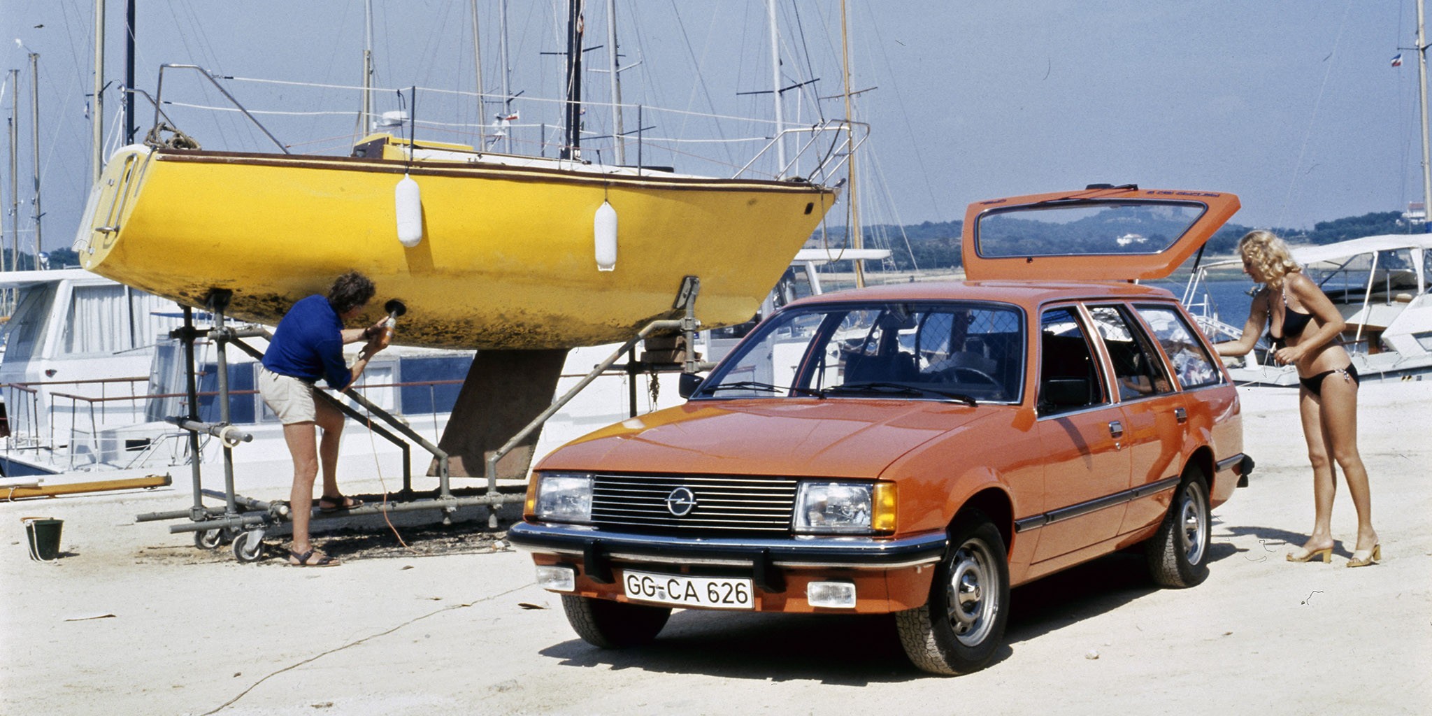 The picture shows a red Opel Rekord E station wagon with the tailgate open next to a yellow boat on the beach and a lady in a bikini.
