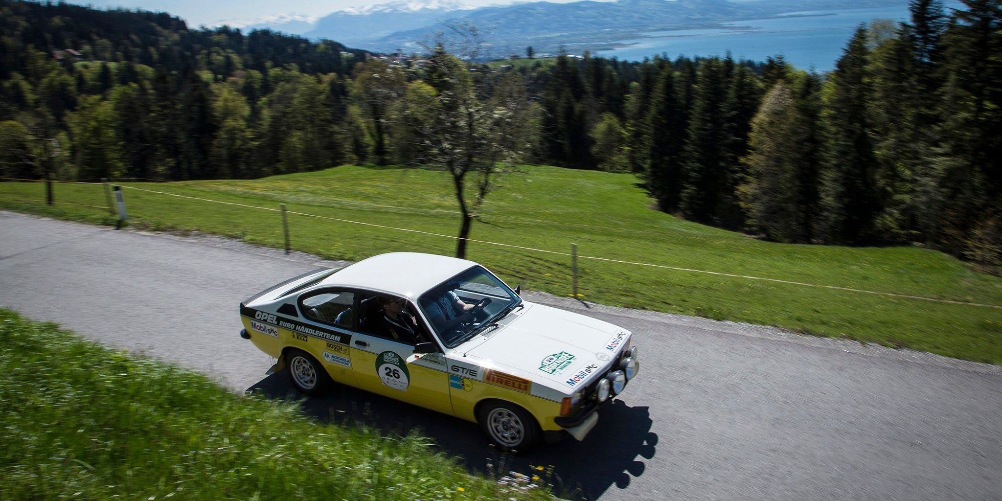 A yellow and white Opel Kadett GT-E coming from the left on a country road. Lake Constance can be seen in the background.