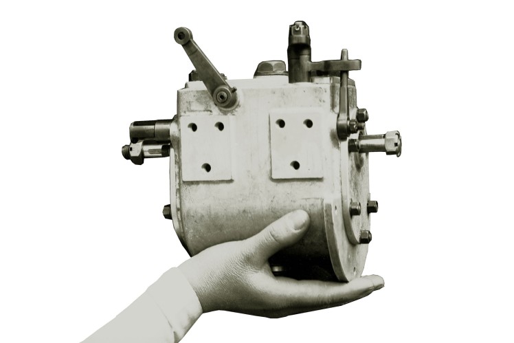 Auf einem Bild in schwarz-weiss hält eine Hand ein altes 3-Gang-Getriebe hoch.