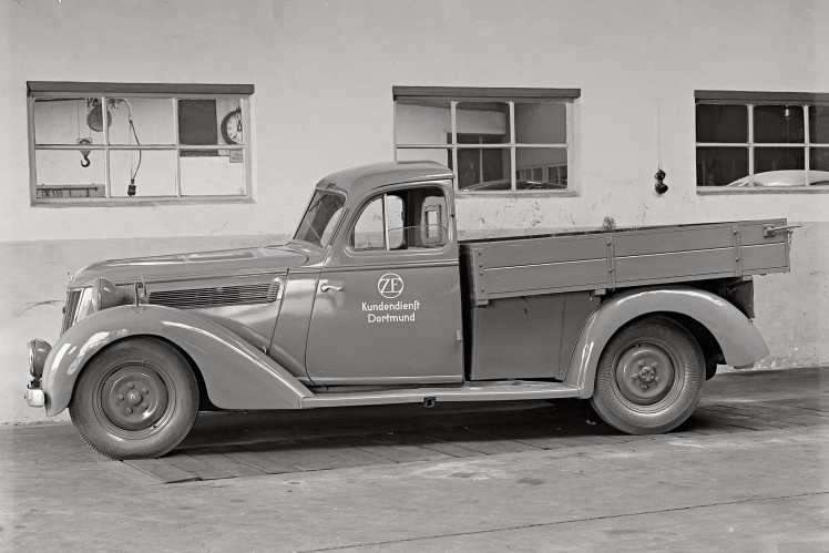 Vor einer Halle steht in schwarz-weiss ein alter Pickup-Kundendienstwagen mit ZF-Logo.
