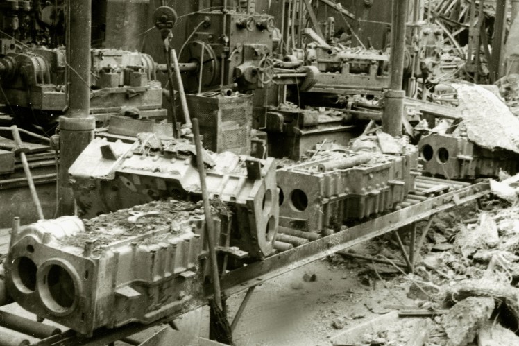 Das Bild in schwarz-weiss zeigt mehrere zerstörte Getriebe auf einem Montageband in einer verwüsteten Halle.