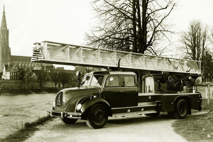 Auf dem Bild in schwarz-weiss ist ein Magirus-Feuerwehrauto von der Seite an einem Fluss zu sehen.