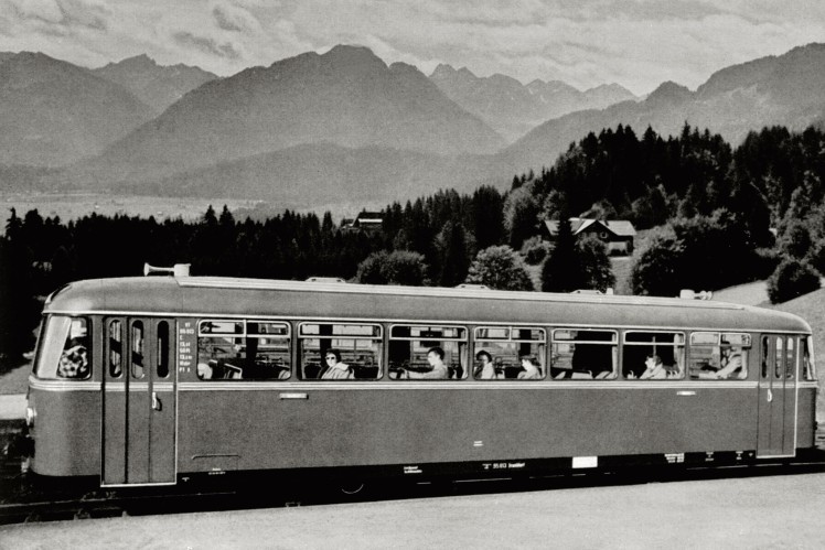 Auf einem Bild in schwarz-weiss ist ein langer Schienenbus von der Seite vor einer bergigen Landschaft zu sehen.
