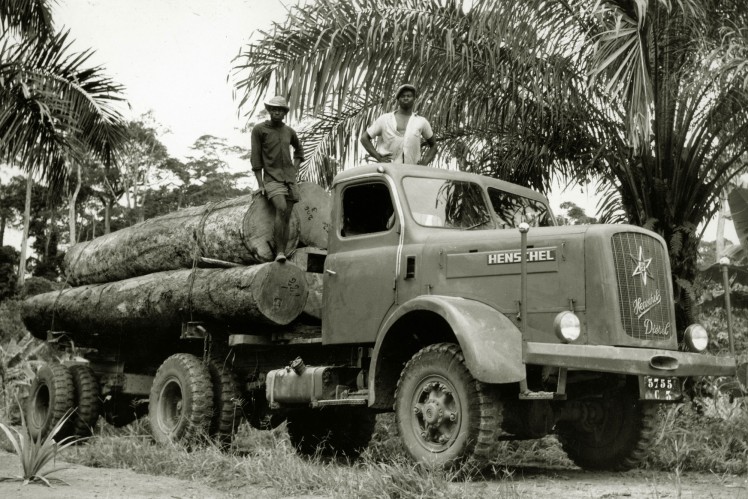 Auf einem Bild in schwarz-weiss ist ein Henschel-Lkw als Holztransporter mit zwei stehenden Männern darauf in den Tropen zu sehen.