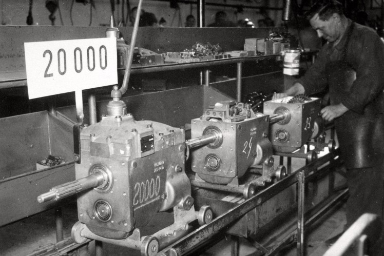 Auf einem Bild in schwarz-weiss sind drei Getriebe auf einem Montageband mit einem ZF-Mitarbeiter und einem Schild zu sehen, auf dem 20000 steht.