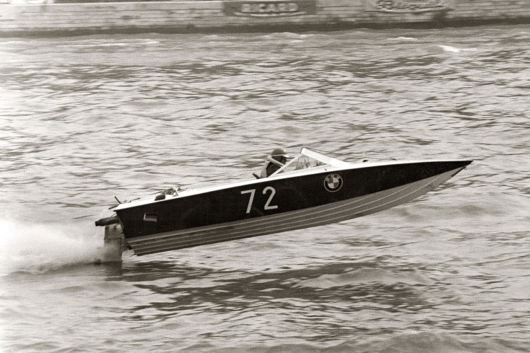 Auf einem Bild in schwarz-weiss ist ein Rennsportboot von der Seite zu sehen, welches über dem Wasser abgehoben ist.