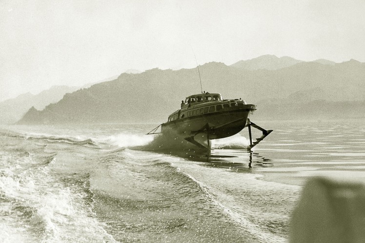 Auf einem Bild in schwarz-weiss ist ein fahrendes Tragflügelboot von vorne auf dem Wasser zu sehen.