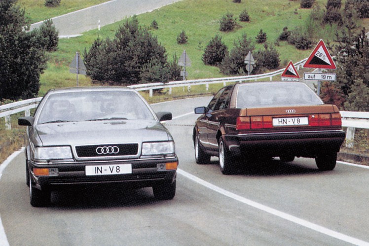 Das Farbbild zeigt nebeneinander zwei Audi V8 jeweils von vorne und hinten, die sich auf einer Strasse begegnen.