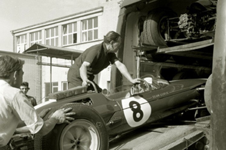Black and white image of lotus car being loaded to transportation by two people.
