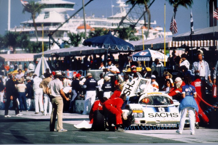 White car on race track during wheel change and spectators viewing.