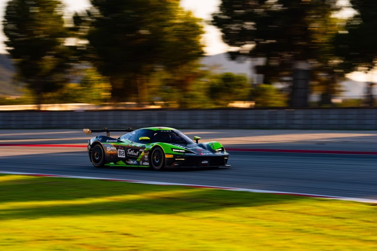 Green and blue race car on race track with blurred trees and grass in background.