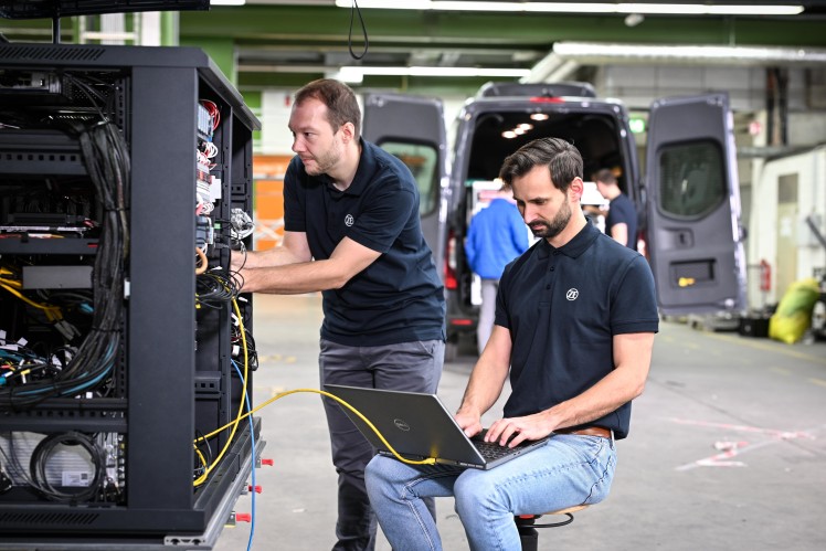 One man sitting on a chair works on a laptop. Another man looks into the trunk of a van, where the car's central computing unit can bee seen.